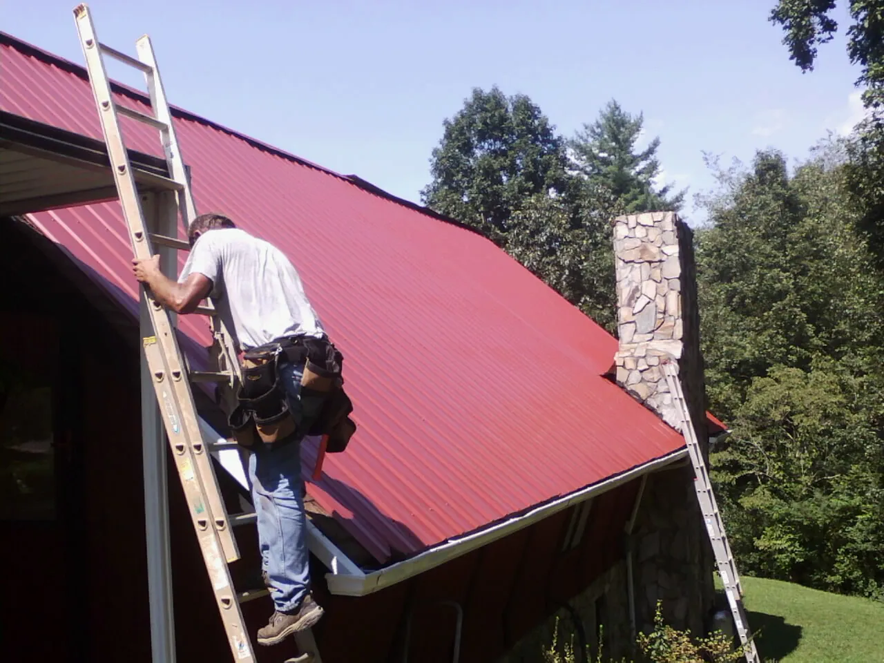 Roofer on a ladder beside a red metal roof and stone chimney — Soffit & Fascia Repair by Roofing Brothers in Hollywood